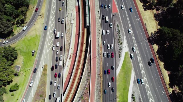 Aerial view of cars navigate through a complex highway interchange while a train moves alongside the busy road in Perth city under clear skies.