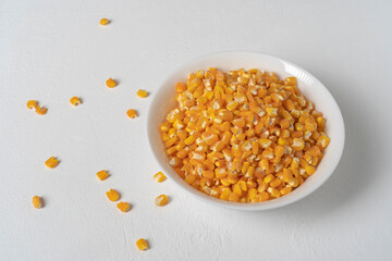 White ceramic bowl.Inside, threshed yellow corn.On a white table.Around it, loose kernels of corn.