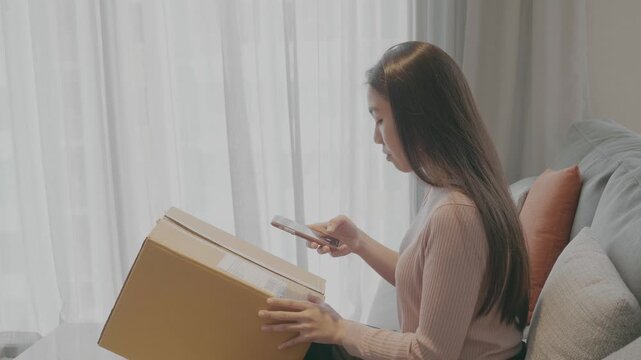 A woman eagerly unpacks a box while checking her phone, embodying excitement and curiosity. The soft natural light enhances the scene, creating a cozy atmosphere.