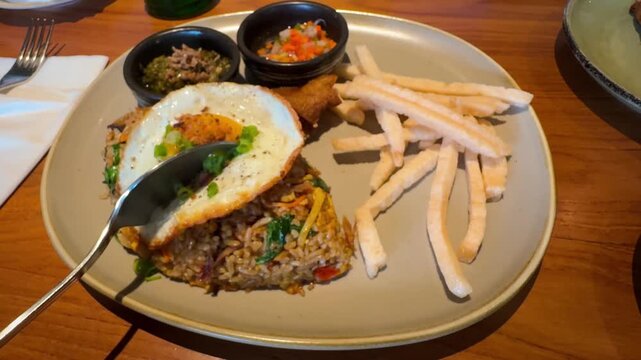 Close-up footage of Indonesian nasi goreng with fried egg, kerupuk sticks, and side sambal on a beige plate, served on a wooden table in a restaurant setting.

