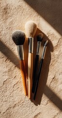 Four makeup brushes with varying bristle types and handle materials, arranged on a textured beige surface, illuminated by a diagonal sunbeam