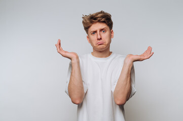 Young man in white t-shirt shrugging with confused expression against plain background.
