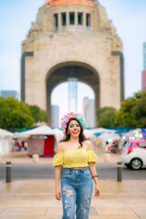 Fototapeta premium Smiling young Latina with colorful catrina makeup walks at Monumento a la Revolucion in Mexico City during Dia de Muertos, wearing yellow top and ripped blue jeans, radiating confidence and tradition