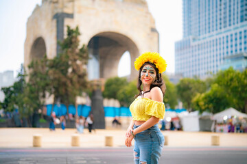 Fototapeta premium Smiling young Latina with colorful catrina makeup walks at Monumento a la Revolucion in Mexico City during Dia de Muertos, wearing yellow top and ripped blue jeans, radiating confidence and tradition