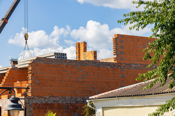 Workers are constructing a new brick building with a crane lifting materials against a backdrop of fluffy clouds and bright sun