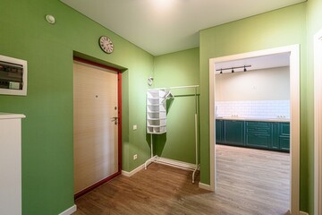 hallway with green walls, a light wood door with a red frame, and a glimpse into a kitchen. Wooden flooring and a white clothing rack are also visible