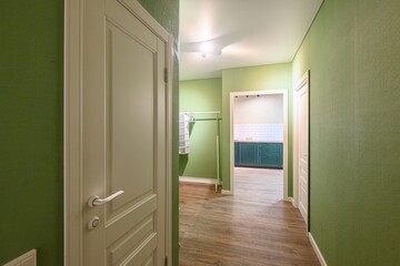 hallway painted in shades of green with wooden floors. A cream-colored door is on the left. In the distance, one can see a kitchen with dark green cabinets and white subway tiles
