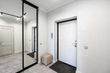 bright entryway features a full-length mirror closet, a white door with black trim, light gray walls, a ceiling light fixture, and a small beige ottoman, creating a clean, modern aesthetic