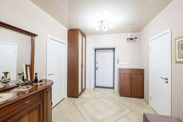 well-lit hallway featuring a wooden dresser with a mirror, a wardrobe, and two white doors. The floor is tiled with a decorative pattern. A white front door is centered in the view