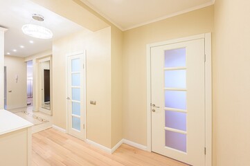 Bright hallway with two white doors with glass panels, light wooden floor, and beige walls. A chandelier illuminates the space