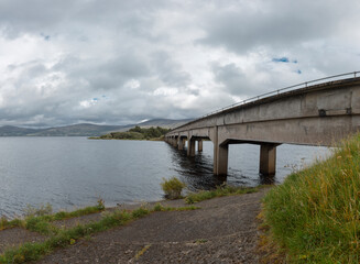 Obraz premium Concrete bridge spanning water with visible shoreline and grassy bank. Irish highland infrastructure crossing with natural landscape and mountain views.
