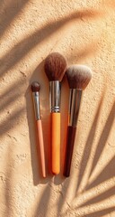 Three makeup brushes with orange and brown handles rest on a beige textured surface, dappled with palm leaf shadows from sunlight