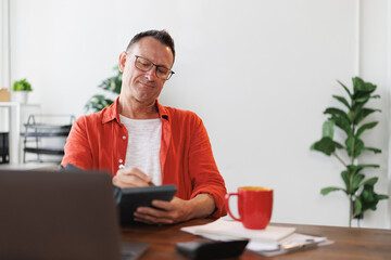 Mature businessman wearing glasses and casual clothes using calculator and taking notes in notebook while working remotely from home office with laptop
