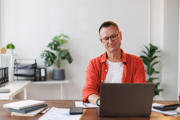 Fototapeta premium Mature businessman with eyeglasses and red shirt sitting at desk and working with laptop in his home office, he is smiling and looking at the screen