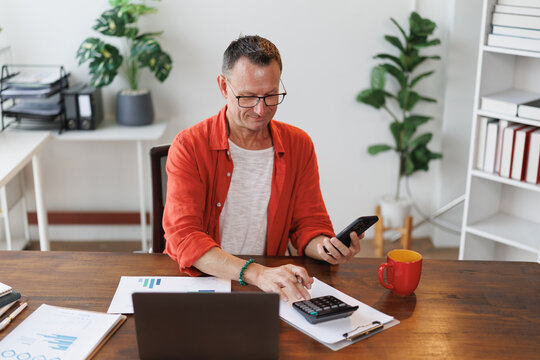 Mature entrepreneur sitting at his desk at home office calculating domestic finances using calculator and looking at his smartphone to check expenses