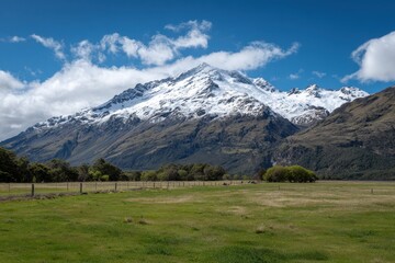Majestic snow capped mountain peak under a vibrant blue sky with scattered fluffy clouds above a green meadow