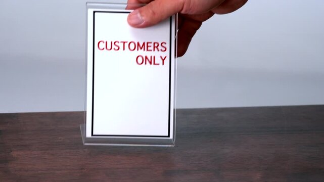 A hand delicately lifts a Customers Only sign from a polished wooden table, as if preparing to place or move it in a reserved space.