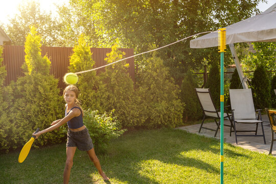 Young girl actively playing tetherball with yellow racket in sun-drenched backyard, enjoying fun summer day outdoors