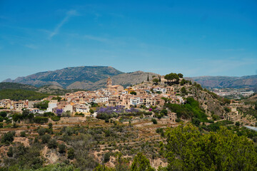 Obraz premium A charming view of Polop, Alicante, a Mediterranean village on a hill with traditional terracotta-roofed houses, surrounded by green trees and mountains under a clear blue sky.