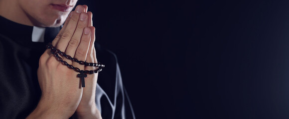 Priest with rosary praying on dark background, closeup. Space for text