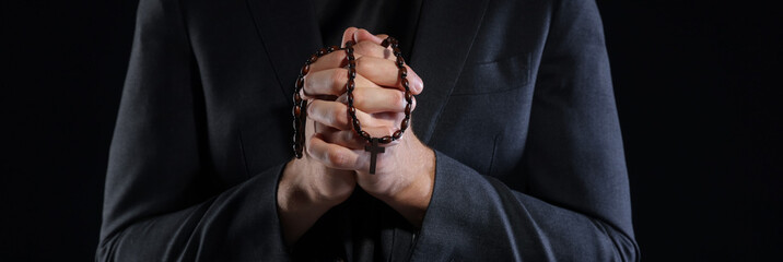 Priest with rosary praying on black background, closeup
