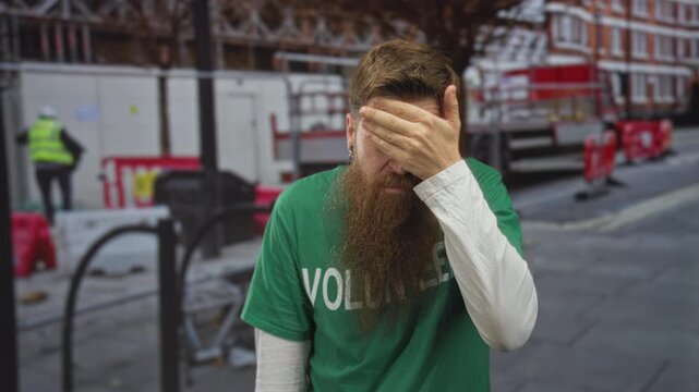 Young bearded man covers face with hand while standing before building scaffolding on street; embarrassment.