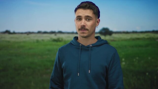 Young man in hoodie stands outdoors in a park with green field background under clear sky exuding casual confidence and relaxed vibe in a serene natural setting