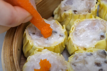 Close-up of siu mai dumplings being topped with bright orange fish roe in a bamboo steamer, highlighting dim sum preparation.