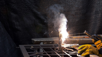 Bright welding sparks and dense smoke rising as gloved hands perform arc welding on metal rods with a black backdrop
