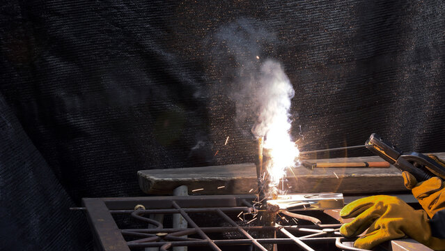 Bright welding sparks and dense smoke rising as gloved hands perform arc welding on metal rods with a black backdrop