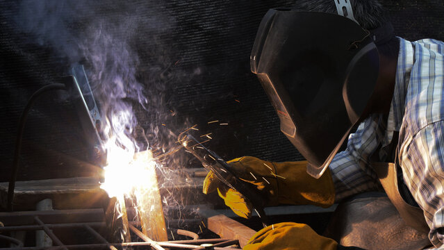 Close-up of focused welder wearing protective helmet and gloves as sparks and thick smoke rise from intense arc welding on metal frame