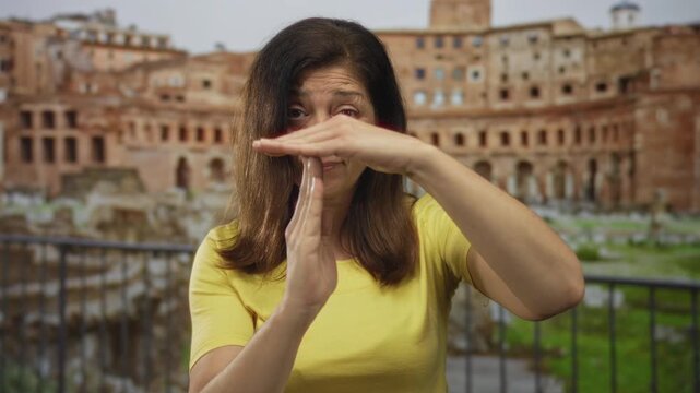 Woman wearing yellow t shirt holds hands in timeout gesture at building railing with roman ruins in view; patience.