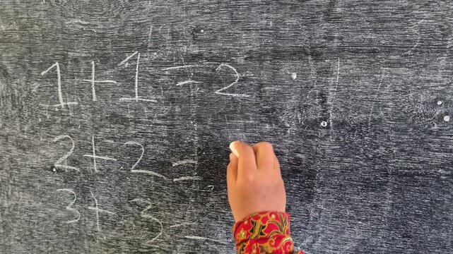 An elementary school student's hand is writing numbers to answer a math problem on the blackboard using chalk.