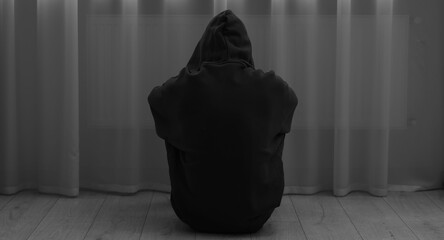 Sadness and depression. Silhouette of man in hoodie near curtains at home, back view. Black and white effect