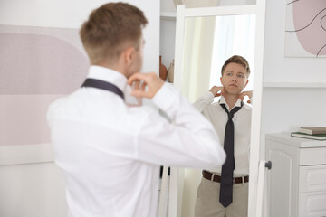 Fototapeta premium Getting ready. Young man tying stylish necktie near mirror at home
