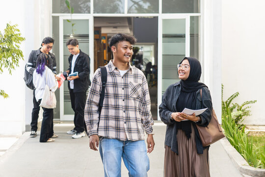 Two Asian students walking outside university building, smiling and chatting after class. Muslim woman and Indonesian man at campus