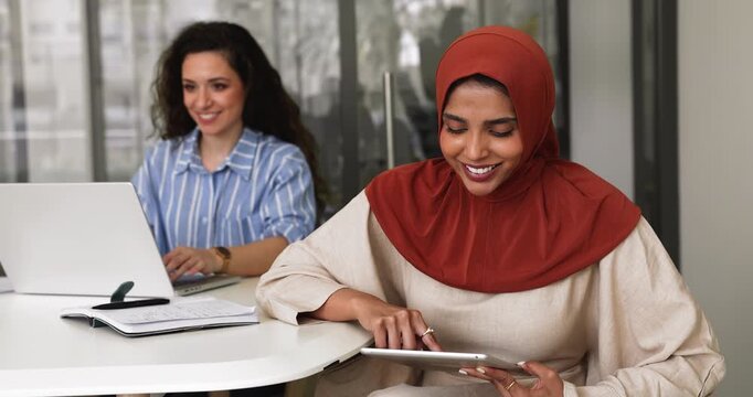 Two multicultural women working on joint task using diverse devices seated in modern office, reviewing document, researching information, or emailing. Digital tools usage in daily business routines - Powered by Adobe