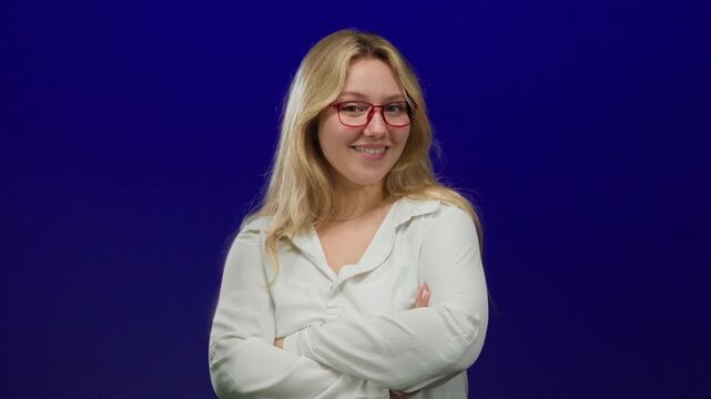 Woman smiling with arms crossed against isolated blue background wearing red glasses and white shirt, exuding confidence and positivity in a studio style setting