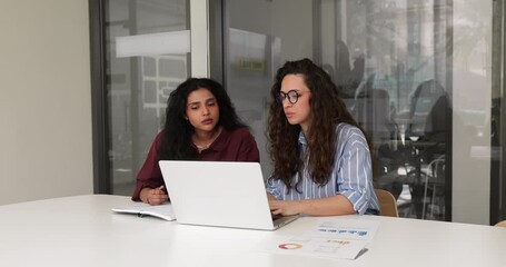 Young 30s European woman conducting meeting, provide explanation data to Indian female colleague or apprentice seated at office desk with papers and laptop. Apprenticeship, coordination, mentorship - Powered by Adobe
