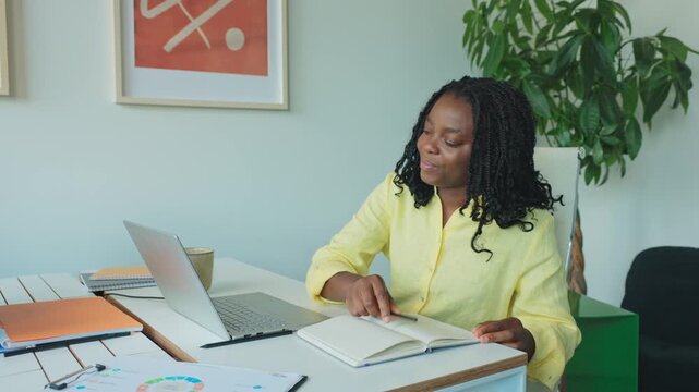 Confident African American woman sitting at white desk taking notes in notebook while glancing at laptop. Planning new task or organizing schedule. Staying productive in calm office environment.