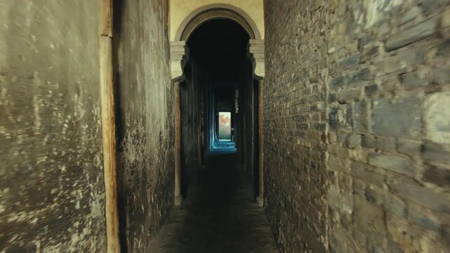 A narrow corridor with a stone archway leading to a distant view of a building. The walls are made of rough, weathered bricks, and the floor appears to be made of cobblestone.