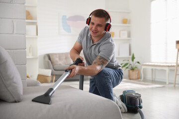 Man in headphones cleaning sofa with vacuum cleaner at home