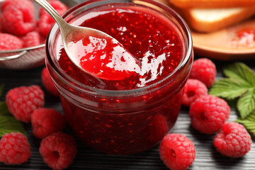 Taking sweet raspberry jam with spoon from glass jar on dark gray table, closeup