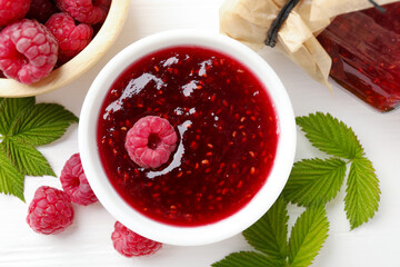 Sweet raspberry jam, berries and green leaves on white wooden table, flat lay