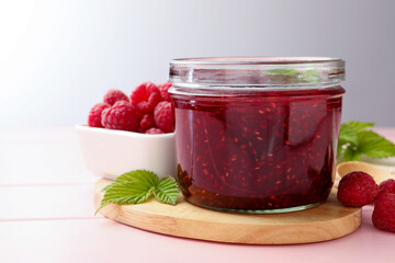 Sweet raspberry jam in glass jar, berries and mint on pink wooden table, closeup