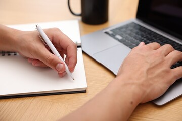 Copywriter using laptop and writing in notebook at wooden table indoors, closeup