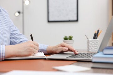 Copywriter using laptop and writing in notebook at wooden table indoors, closeup
