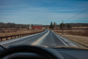 View from car interior showing curved country road leading to red barn and farm buildings in rural countryside setting