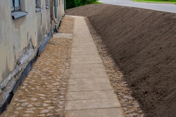 Narrow sidewalk along an old building under renovation. Freshly leveled soil borders one side of the path, while the other side shows a weathered wall with peeling paint and stone edging.
