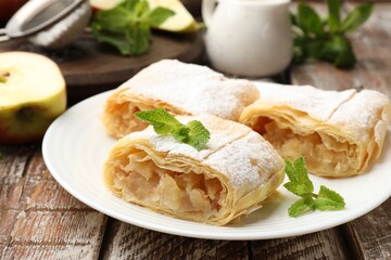 Pieces of tasty apple strudel with powdered sugar and mint on wooden table, closeup